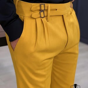May include: Mustard yellow trousers with a double-buckle belt and pleated front. The trousers are paired with a black shirt. The image includes the text "Look Iconic with REALEVER" and the REALEVER logo.