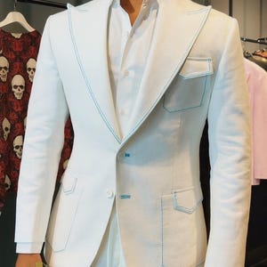 May include: White suit with light blue stitching detail. The suit features a tailored jacket with flap pockets and a matching pair of trousers. The jacket has a notched lapel and a two-button closure. The suit is made of a lightweight fabric.