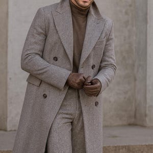 May include: A man's ensemble featuring a brown fedora, a brown turtleneck, and a double-breasted gray coat. The outfit includes matching gray trousers and brown leather gloves. The text "Look Iconic with REALEVER" is visible.