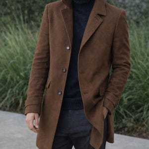 May include: A brown overcoat with a notched lapel and three buttons. The coat is worn over a navy blue turtleneck sweater and dark gray pants. The image includes the text "REALLEVER".
