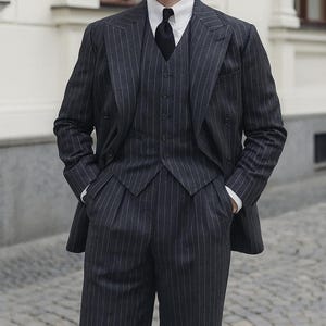 May include: A three-piece charcoal gray pinstripe suit with a white dress shirt and black tie. The suit includes a double-breasted jacket, vest, and pleated trousers. The text "Look Iconic with REALEVER" is visible.