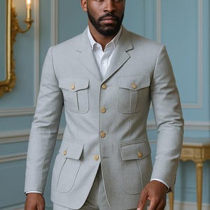 May include: A light grey suit comprising a jacket and trousers. The jacket features four front pockets and gold-coloured buttons. The suit is worn over a white dress shirt. The suit is a formal wear item.