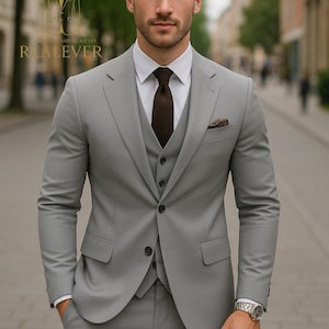 May include: A light gray three-piece suit with a white dress shirt and a brown tie. The suit includes a jacket, vest, and trousers. A pocket square and a wristwatch complete the ensemble. The suit is tailored and worn outdoors.