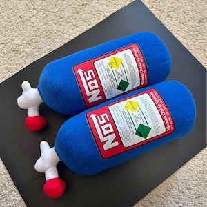 May include: Two blue plush cylindrical pillows shaped like nitrous oxide tanks. Each features a red and white label with the text NOS, Niteous Oxide Systems, and warning symbols. The pillows have white valve handles with red tips at one end.