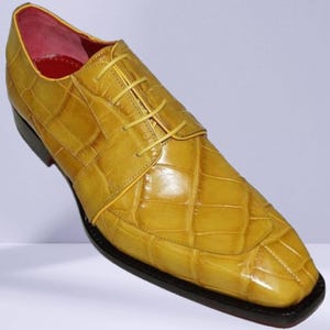 May include: A single yellow leather dress shoe with a textured, crocodile-like pattern. The shoe has a square toe, a black sole, and yellow laces. The interior is lined with red leather.