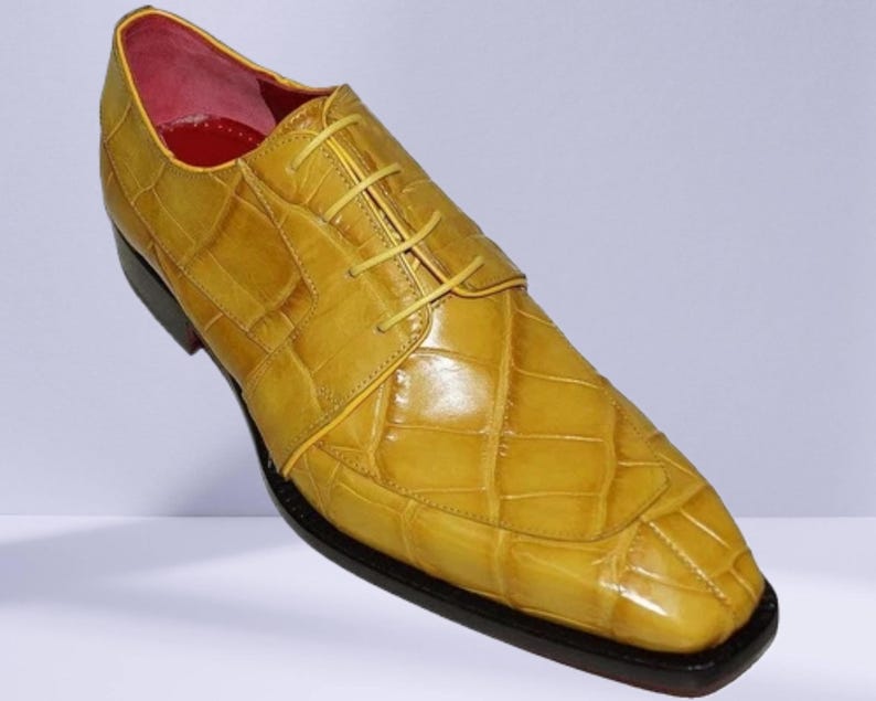 May include: A single yellow leather dress shoe with a textured, crocodile-like pattern. The shoe has a square toe, a black sole, and yellow laces. The interior is lined with red leather.