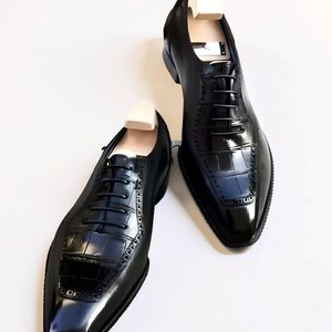 Peut inclure: Une paire de chaussures richelieu en cuir noir avec une finition brillante et un motif gaufré façon crocodile sur l'empeigne. Les chaussures ont des lacets noirs, un bout pointu et des embauchoirs en bois clair insérés dans chaque chaussure.