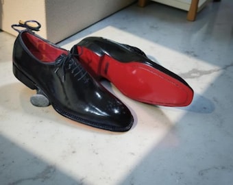 May include: A pair of black leather dress shoes with a glossy finish. The shoes have red soles and red interior lining. The shoes are laced up and are sitting on a light-colored surface.