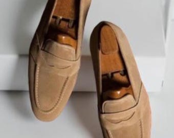 May include: Two tan suede penny loafers with wooden shoe trees. The shoes have a classic design with a rounded toe and a strap across the vamp. The loafers are displayed on a white surface.