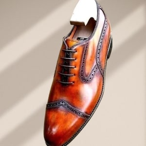 May include: A single brown leather dress shoe with a wooden shoe tree inserted. The shoe features a cap toe design with decorative perforations and a lace-up closure. The leather has a polished, glossy finish.