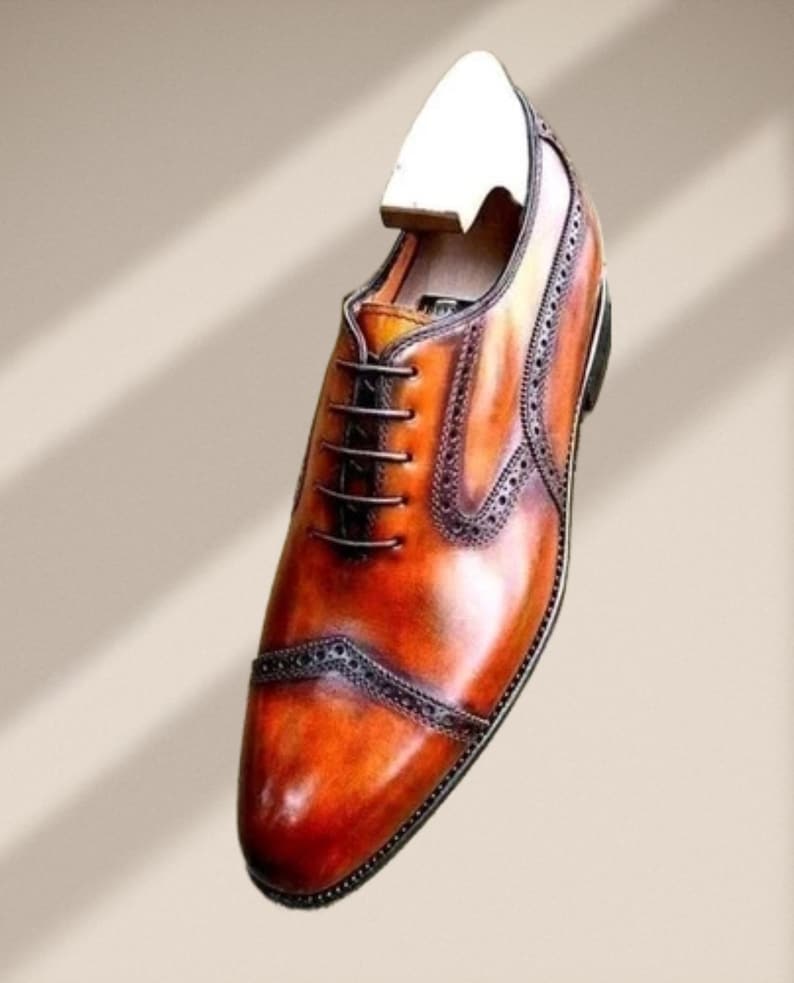 May include: A single brown leather dress shoe with a wooden shoe tree inserted. The shoe features a cap toe design with decorative perforations and a lace-up closure. The leather has a polished, glossy finish.