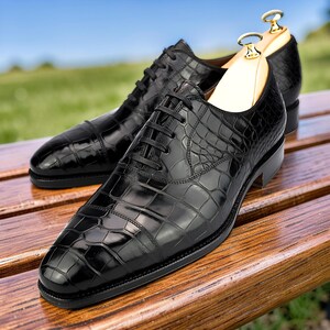 Peut inclure: Une paire de chaussures oxford noires présentant une texture brillante en relief façon crocodile. Les chaussures sont dotées d'un système de laçage fermé avec des lacets noirs fins et incluent des embauchoirs en bois clair pour garder la forme.