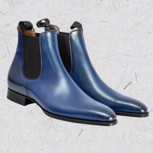 Puede incluir: Un par de botines Chelsea azules con un acabado brillante. Los botines tienen punta redonda, paneles laterales elásticos y una lengüeta en la parte trasera. Las suelas y los ribetes son negros. Los botines son de cuero.
