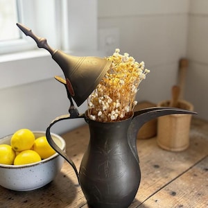 May include: A dark metal coffee pot with a long spout and handle, filled with dried yellow flowers. A matching lid is tilted to the side. A bowl of lemons sits nearby on a wooden table, creating a rustic, decorative display.
