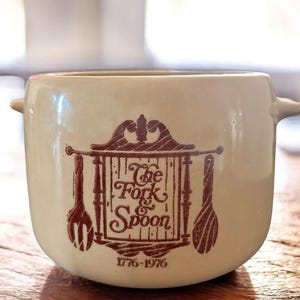 Peut inclure: Bol en céramique crème avec deux poignées. Le bol présente un motif marron d'une fourchette et d'une cuillère avec le texte "The Fork & Spoon" et les dates "1776-1976". Ce bol vintage est adapté pour servir de la soupe ou d'autres plats.