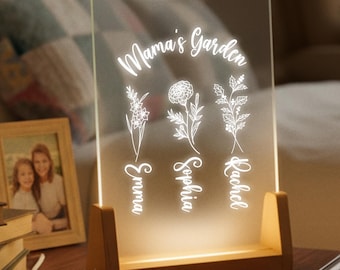 May include: A lighted acrylic sign with the words "Mama's Garden" and the names Emma, Sophia, and Rachel. The sign features floral designs and sits in a wooden base. A framed photo and books are on the nightstand.