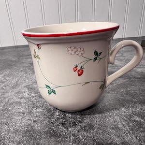 Puede incluir: Una taza de cerámica de color crema con un delicado patrón botánico de pequeñas fresas rojas, hojas verdes y flores blancas. La taza tiene un borde ancho y acampanado acentuado con una fina línea roja pintada y un asa curva a la derecha.