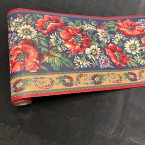 May include: A roll of floral wallpaper featuring a repeating pattern of large red poppies, white daisies, and green leaves on a navy blue background. The border has a tan and green decorative trim with a red edge.