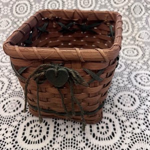 Puede incluir: Una pequeña cesta cuadrada tejida en tonos marrones, con detalles en verde oscuro. La cesta presenta un adorno en forma de corazón y lazos verdes decorativos. La cesta probablemente se utiliza para almacenamiento o decoración.