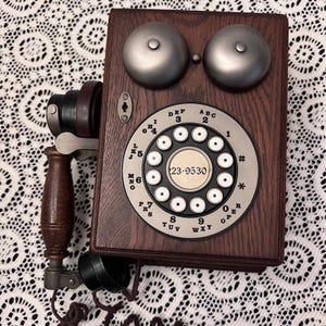 May include: A vintage wall-mounted telephone with a dark wood finish. It features a rotary dial with numbers and letters, two silver bells, and a handset. The phone's dial displays the number 23-9530. The design evokes a classic, antique aesthetic.