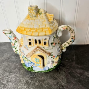 Puede incluir: Una tetera de cerámica con forma de cabaña con techo de tejas amarillas, chimenea y puerta abierta. El exterior blanco presenta enredaderas pintadas, flores coloridas y una pequeña figura de perro cerca de la entrada. El asa y el pico están decorados con motivos florales.