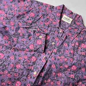 May include: A purple button-down shirt with a floral pattern of pink flowers and green leaves. The shirt has a collar, long sleeves, and wooden buttons. The tag reads "NORTHERN".