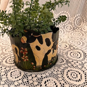 Könnte beinhalten: Ein schwarz-cremefarbener Pflanztopf mit Kuhdesign, gefüllt mit grünen Kunstpflanzen. Der Pflanztopf hat ein handgemaltes Design von Kühen und Blumen. Der Rand des Pflanztopfs hat einen gewellten Rand.