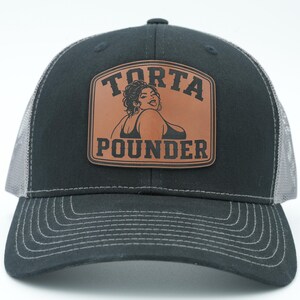May include: A black baseball cap with a mesh back and a brown leather patch on the front. The patch features a graphic illustration of a person and the text "TORTA POUNDER" in black lettering. The cap has a curved brim with visible stitching.