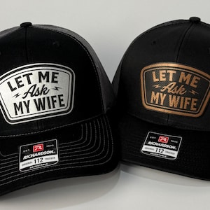 May include: Two black trucker hats with the text "LET ME Ask MY WIFE" on a patch. One patch is white with black text, the other is brown with gold text. Both hats have a black brim and a gray mesh back.