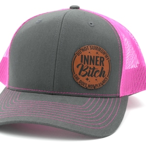 May include: A charcoal gray trucker hat with bright neon pink mesh back panels and matching pink stitching on the brim. A circular brown leather patch is centered on the front, featuring the text DO NOT SUMMON MY INNER BITCH SHE DOES NOT PLAY NICE.