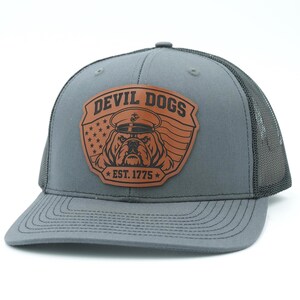 May include: A grey trucker hat with a brown leather patch on the front. The patch features a bulldog wearing a military-style cap, the text "DEVIL DOGS" and "EST. 1775", and a background design of the American flag with stars and stripes.