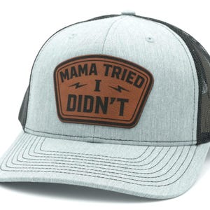 May include: A light gray heathered trucker hat with a black mesh back and a brown leather-like patch on the front. The patch features the black text MAMA TRIED I DIDN'T with two small lightning bolt icons centered between the lines of text.