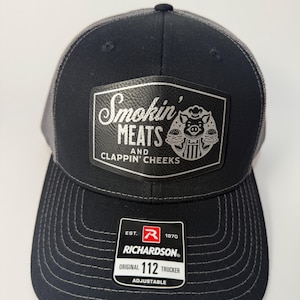 May include: Black and gray trucker hat with a curved brim. The hat features a patch with the text "Smokin' MEATS AND CLAPPIN' CHEEKS" and a pig graphic. The hat also has a Richardson tag that says "ORIGINAL 112 TRUCKER ADJUSTABLE".