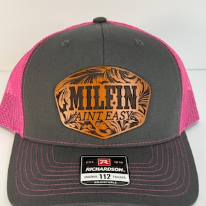 May include: A gray and pink trucker hat with the text "MILFIN AINT EASY" on a brown leather patch. The hat has a pink mesh back and a curved brim with pink stitching. The Richardson brand label is visible.