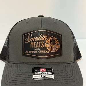 May include: A gray trucker hat with a black mesh back. The front features a rectangular patch with the words "Smokin' MEATS AND CLAPPIN' CHEEKS" and a pig graphic. The hat has a curved brim and an adjustable strap.