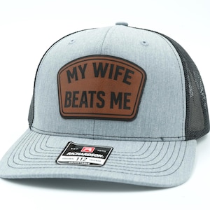 May include: A heather grey trucker hat with a black mesh back and a brown leather-like patch on the front. The patch features the black text MY WIFE BEATS ME. A rectangular Richardson 112 sticker is centered on the flat brim of the baseball cap.
