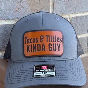 May include: Gray trucker hat with a black mesh back and a brown leather patch. The patch features the text "Tacos & Titties Kinda Guy". The hat has a curved brim and an adjustable strap. The Richardson brand label is visible.