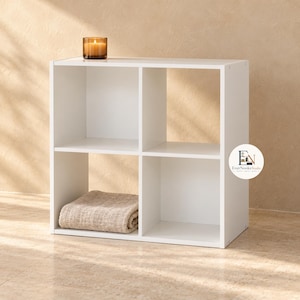 May include: A white four-cube storage organizer with a square frame and a central divider. A lit candle in a glass jar sits on the top surface, and a folded beige textured fabric cloth is placed inside the bottom left storage compartment.