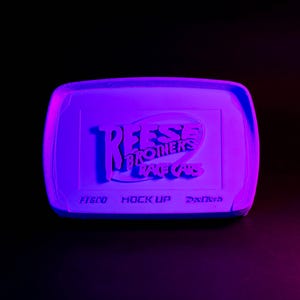 May include: A rectangular, purple-toned object with rounded corners, illuminated against a black background. The text "Reese Brothers Race Cars" is embossed on the surface. Below, smaller text reads "Fisco, Mock Up, Drifted".