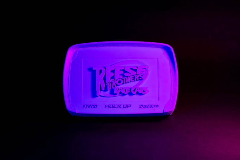 May include: A rectangular, purple-toned object with rounded corners, illuminated against a black background. The text "Reese Brothers Race Cars" is embossed on the surface. Below, smaller text reads "Fisco, Mock Up, Drifted".