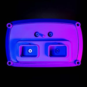 May include: A rectangular, light purple electronic device with rounded edges, featuring two recessed compartments and several circular ports. The device is illuminated with blue and purple lighting against a black background.