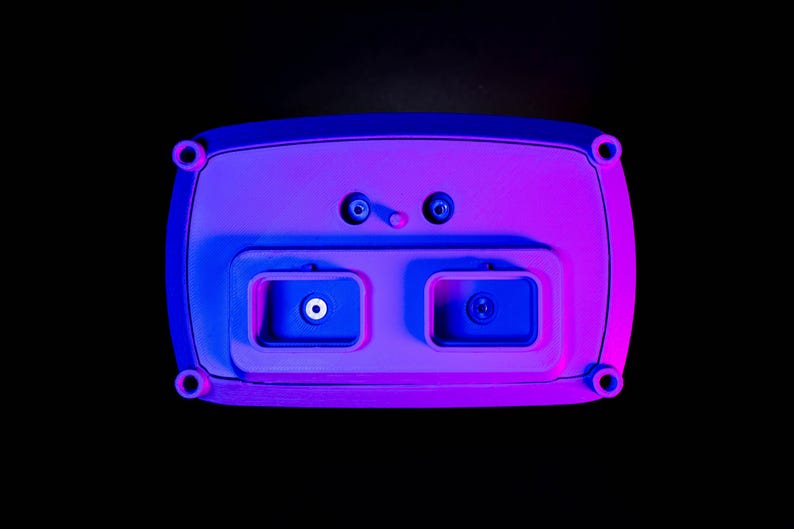 May include: A rectangular, light purple electronic device with rounded edges, featuring two recessed compartments and several circular ports. The device is illuminated with blue and purple lighting against a black background.