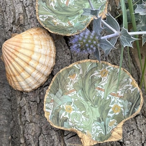 May include: Three decorative dishes in the shape of seashells. One is a natural beige colour, while the other two have a green and white floral pattern with gold trim. They are arranged on a textured, brown surface.