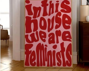 May include: A framed pink poster with the words "In this house we are Feminists" in red, bold, bubble letters. The poster is displayed on a wooden floor, leaning against a white wall.