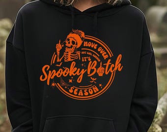 Puede incluir: Sudadera con capucha negra con un gráfico naranja. El gráfico presenta un esqueleto con sombrero haciendo una peineta, con el texto "Move Over Hot Girl Summer It's Spooky B*tch Season" en un diseño circular.