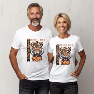 May include: White t-shirts with a tiger graphic and the text "Mitchell High Class of 1985" in orange and black. The shirts also include "40th Class Reunion" text. A man and a woman are seen wearing the matching shirts.