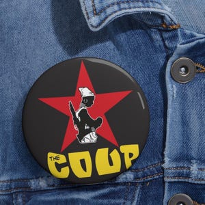May include: A round black button with a red star and a silhouette of a person holding a rifle. The text "THE COUP" is in yellow, with the word "THE" in white. The button is pinned to a denim jacket.