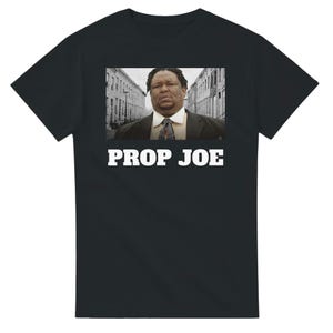 May include: Black t-shirt with a rectangular image of a man in a suit and tie, set against a backdrop of buildings. The text "PROP JOE" is printed in white block letters below the image.