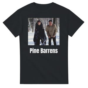 May include: Black t-shirt featuring a rectangular image of two men in a snowy forest. The image is above the white text "Pine Barrens". The men are wearing jackets and standing in the snow.