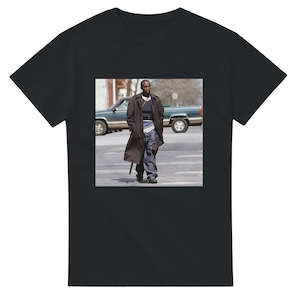 May include: Black t-shirt featuring a photograph of a person walking on a crosswalk. The person is wearing a long brown coat, blue jeans, and a striped shirt. A green pickup truck is in the background.